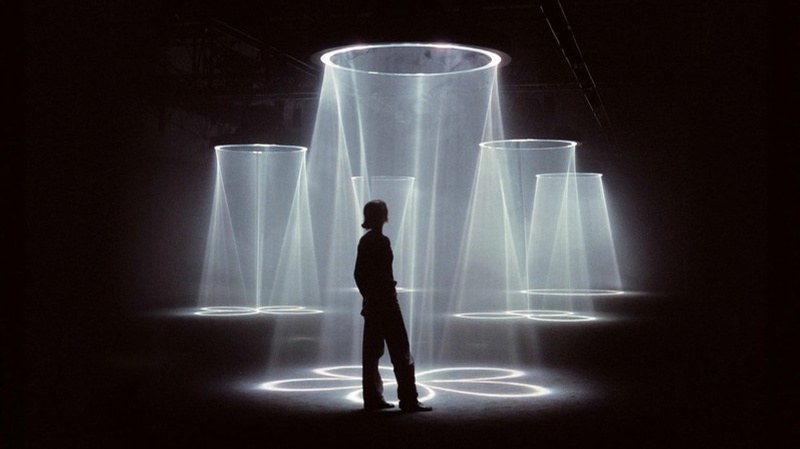 A figure standing among cones of light in a dark room