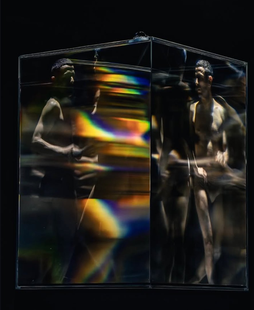 Two figures reflected and refracted inside a glass cube, with bands of rainbow light